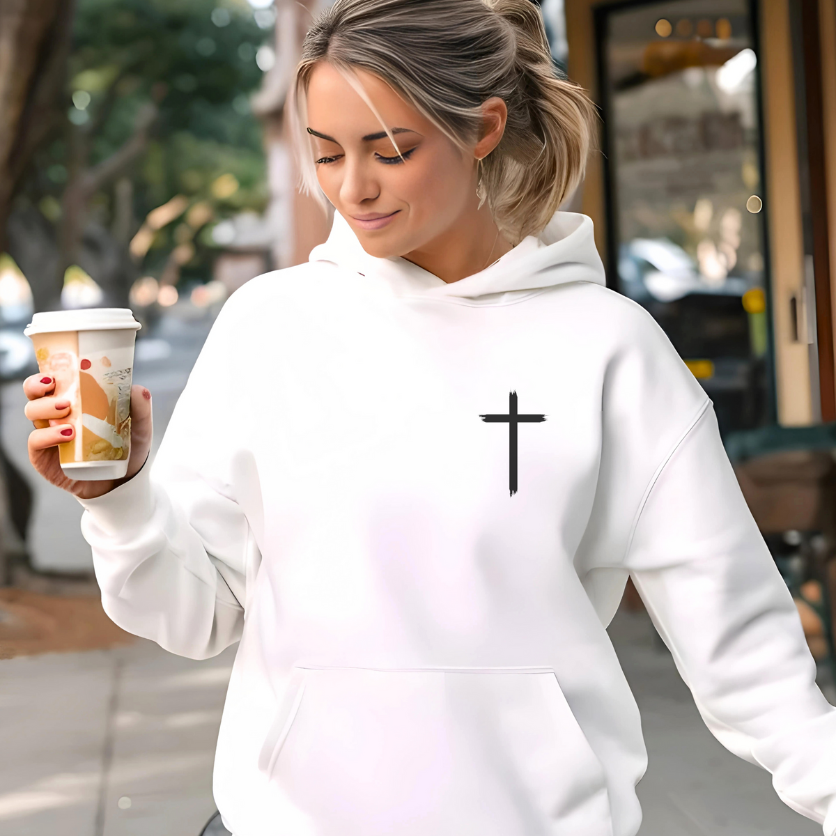 Woman wearing a white hoodie with a black cross on the front chest while holding a coffee cup outdoors.
