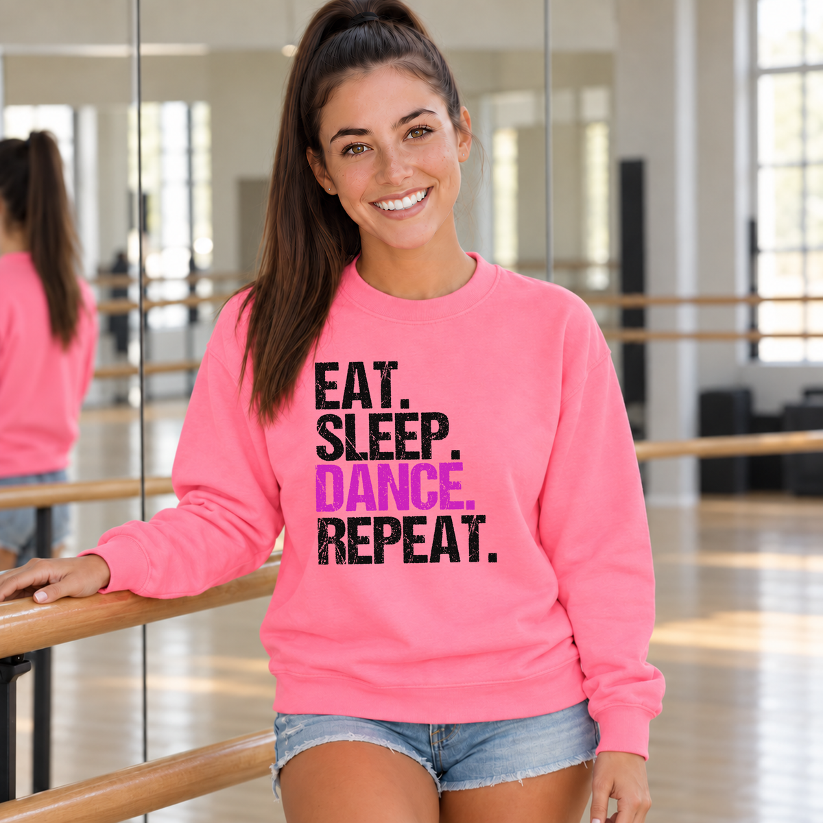 Woman in a bright pink Eat Sleep Dance Repeat sweatshirt beside a dance barre and mirror.