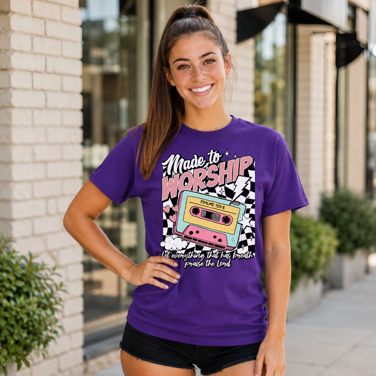 Woman wearing a purple Made to Worship T-shirt with a retro cassette graphic outside a storefront.