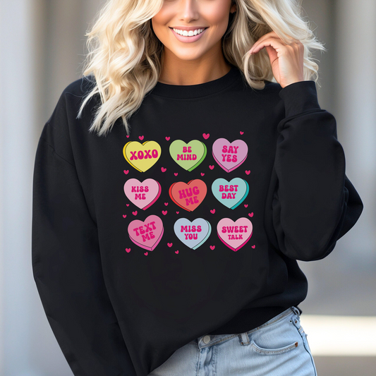 Woman wearing a black Valentine’s sweatshirt featuring colorful candy conversation hearts with messages like “XOXO,” “Be Mine,” “Say Yes,” “Kiss Me,” “Hug Me,” and more.