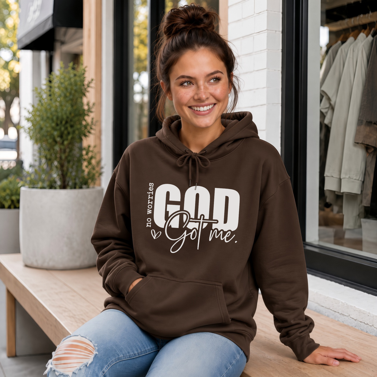 Woman wearing a brown No Worries God Got Me hoodie with bold white graphic lettering outside a boutique.