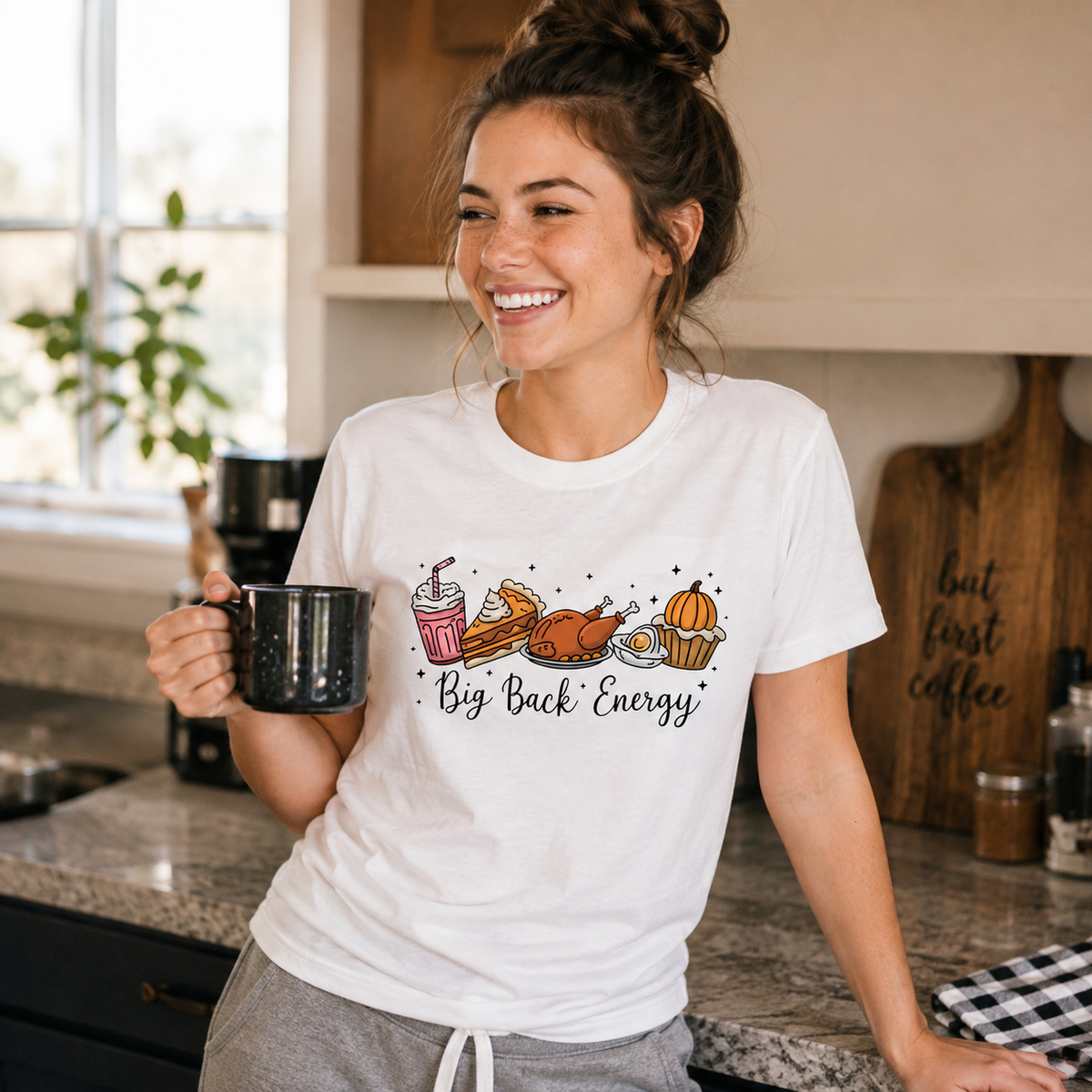 Woman wearing a Big Back Energy Thanksgiving T-shirt holds a mug in a cozy kitchen lifestyle scene.