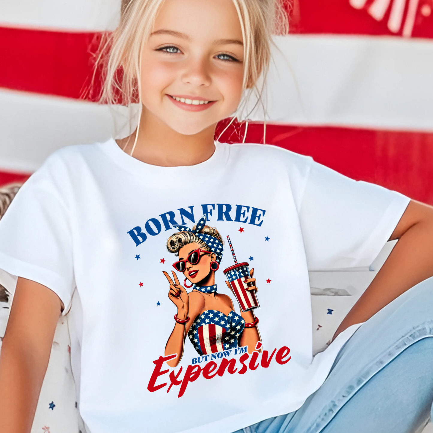 Child wearing a white t-shirt with 'Born Free But Now It's Expensive' graphic design in front of an American flag.