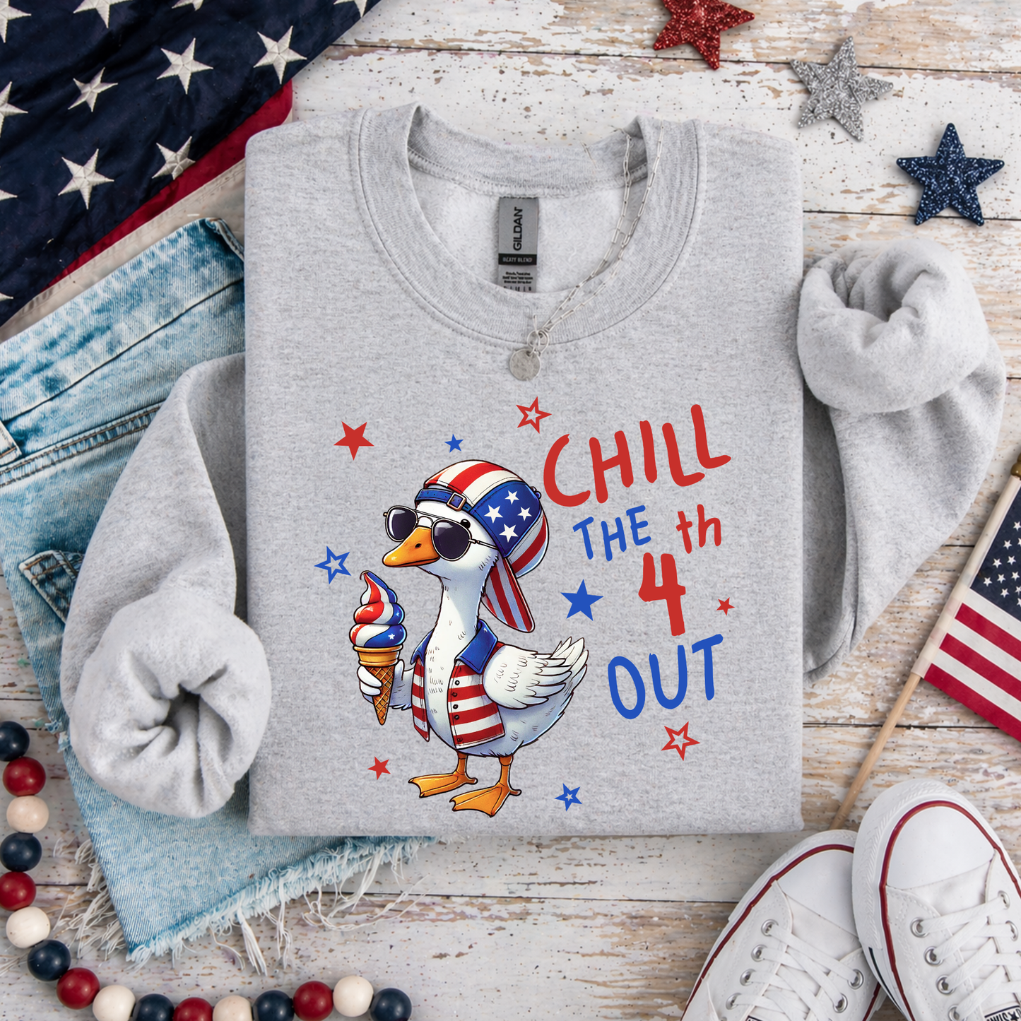 Gray sweatshirt with a duck wearing an American flag hat and ice cream, text 'Chill the 4th Out', on a wooden surface with American flags and jeans.