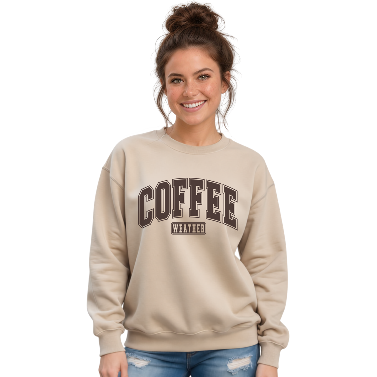 Woman wearing a sand crewneck sweatshirt with "COFFEE WEATHER" printed in dark brown varsity letters, paired with blue jeans against a white background.