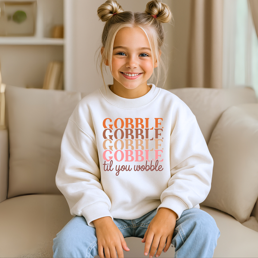 Girl in a white unisex youth sweatshirt with bold “Gobble Til You Wobble” design in orange, pink, and brown. Fall-themed kids’ clothing for Thanksgiving dinner, classroom events, and festive family fun.