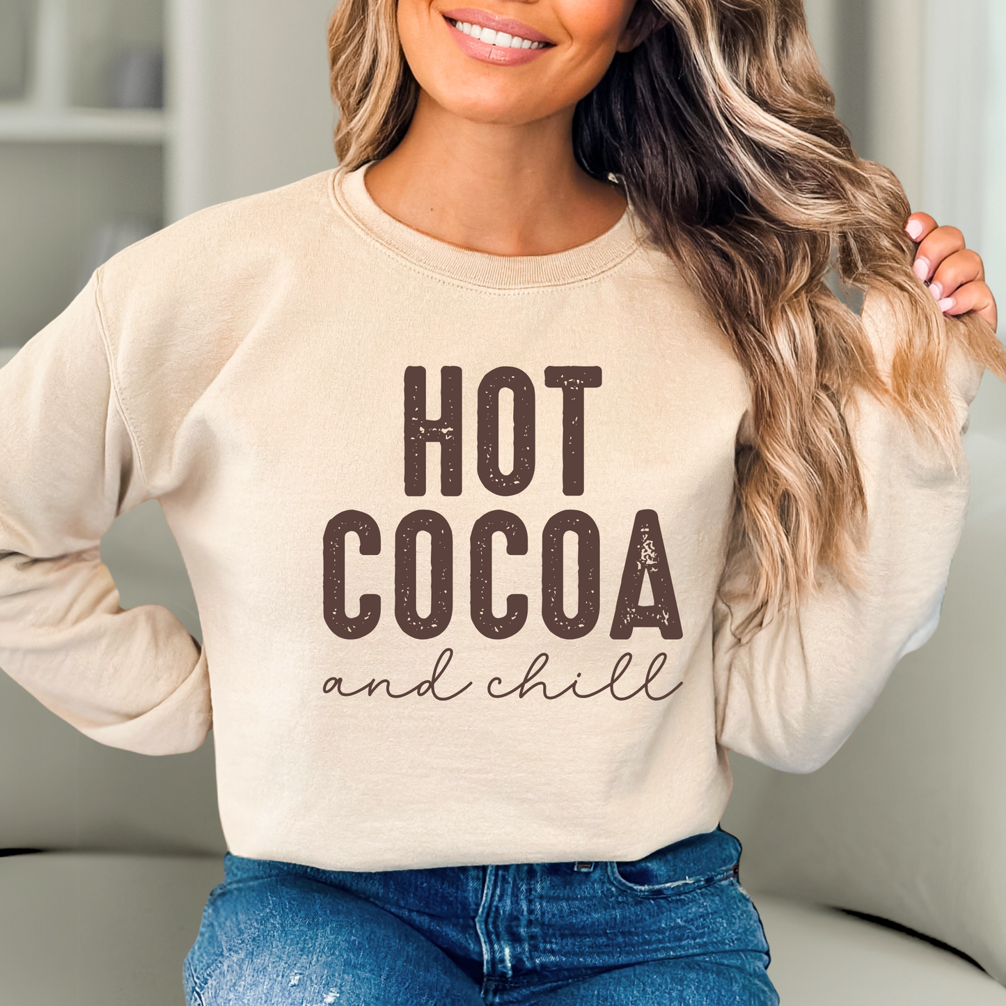 Woman wearing a beige sweatshirt with 'Hot Cocoa and Chill' text in a cozy indoor setting.