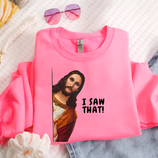 Pink sweatshirt with a graphic of a jesus saying 'I saw that!' text, placed on a textured surface.