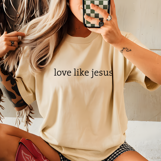 Person wearing a beige t-shirt with 'love like jesus' text, taking a mirror selfie.