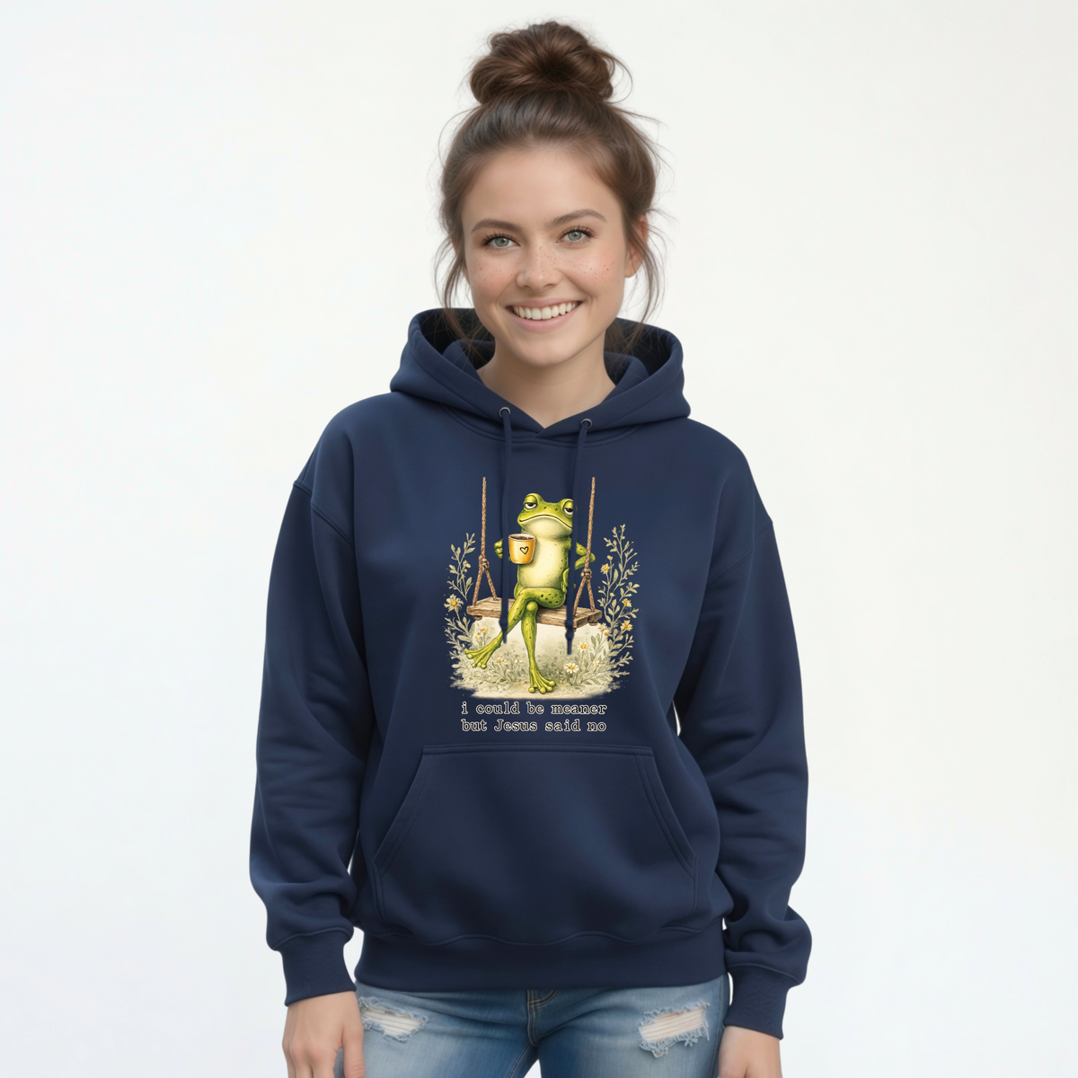 Woman modeling a navy hoodie with a frog sitting on a swing holding a mug and the words “I could be meaner but Jesus said no.”