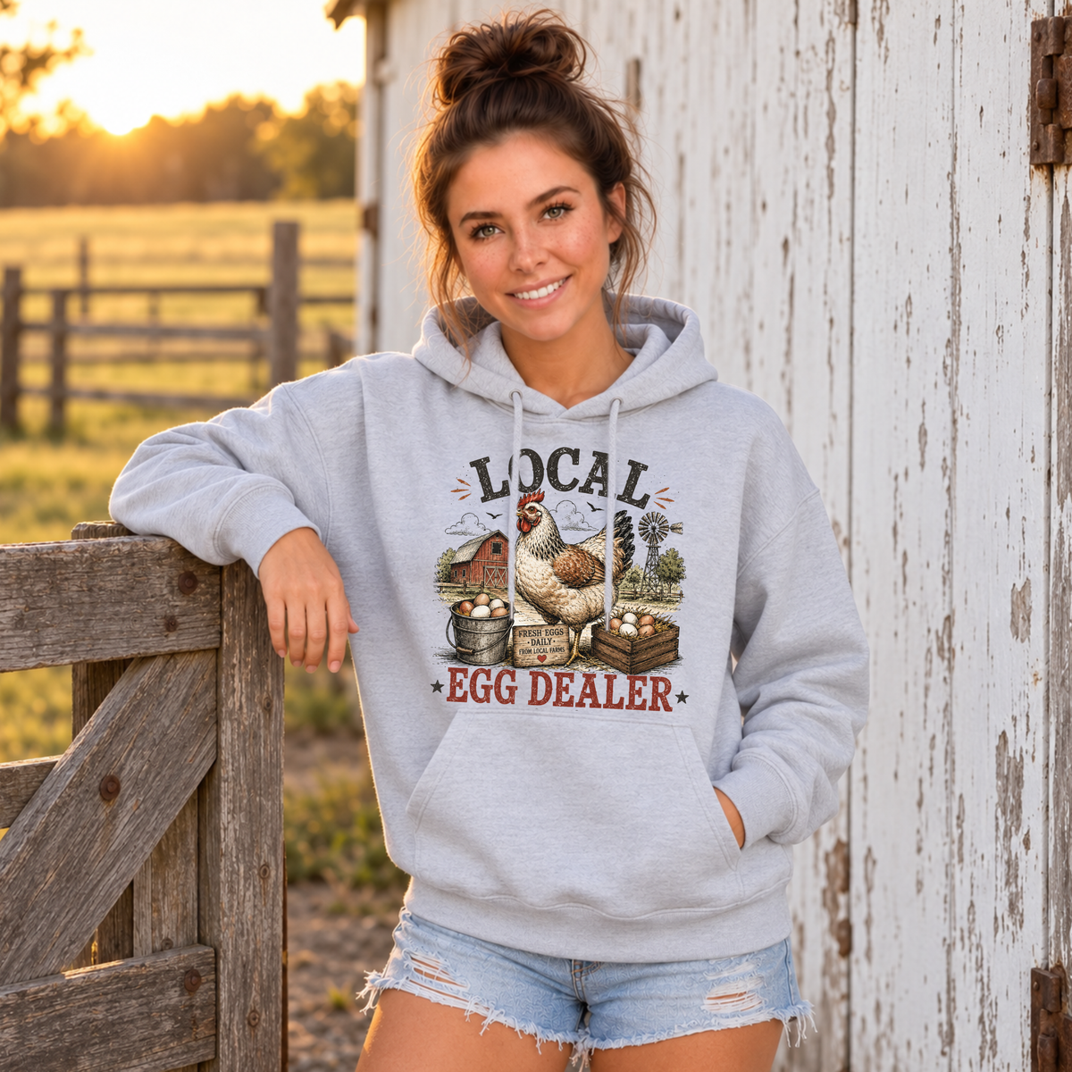Woman in a sports gray Local Egg Dealer hoodie leaning on a farm gate at sunset.