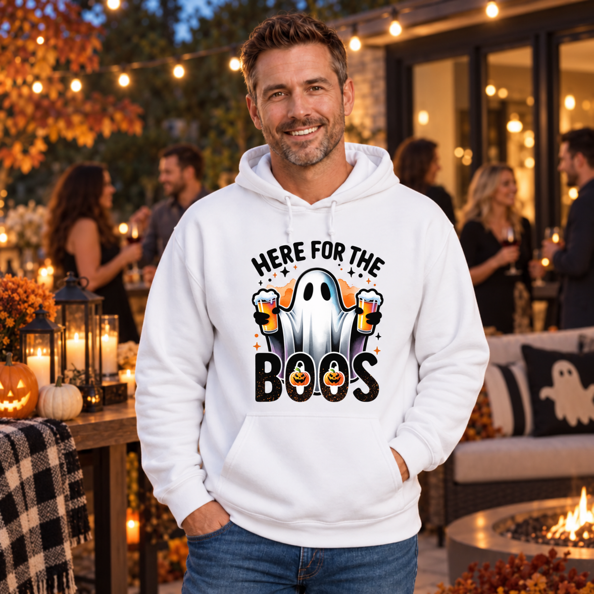 Man wearing a white hoodie featuring a ghost holding two beer mugs and Here For The Boos text at an outdoor Halloween party.