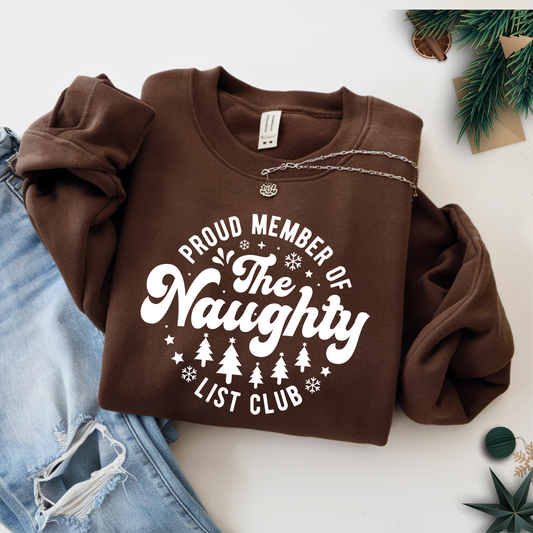 Brown sweatshirt folded flat with necklace and pine décor – “Proud Member of the Naughty List Club” holiday graphic.