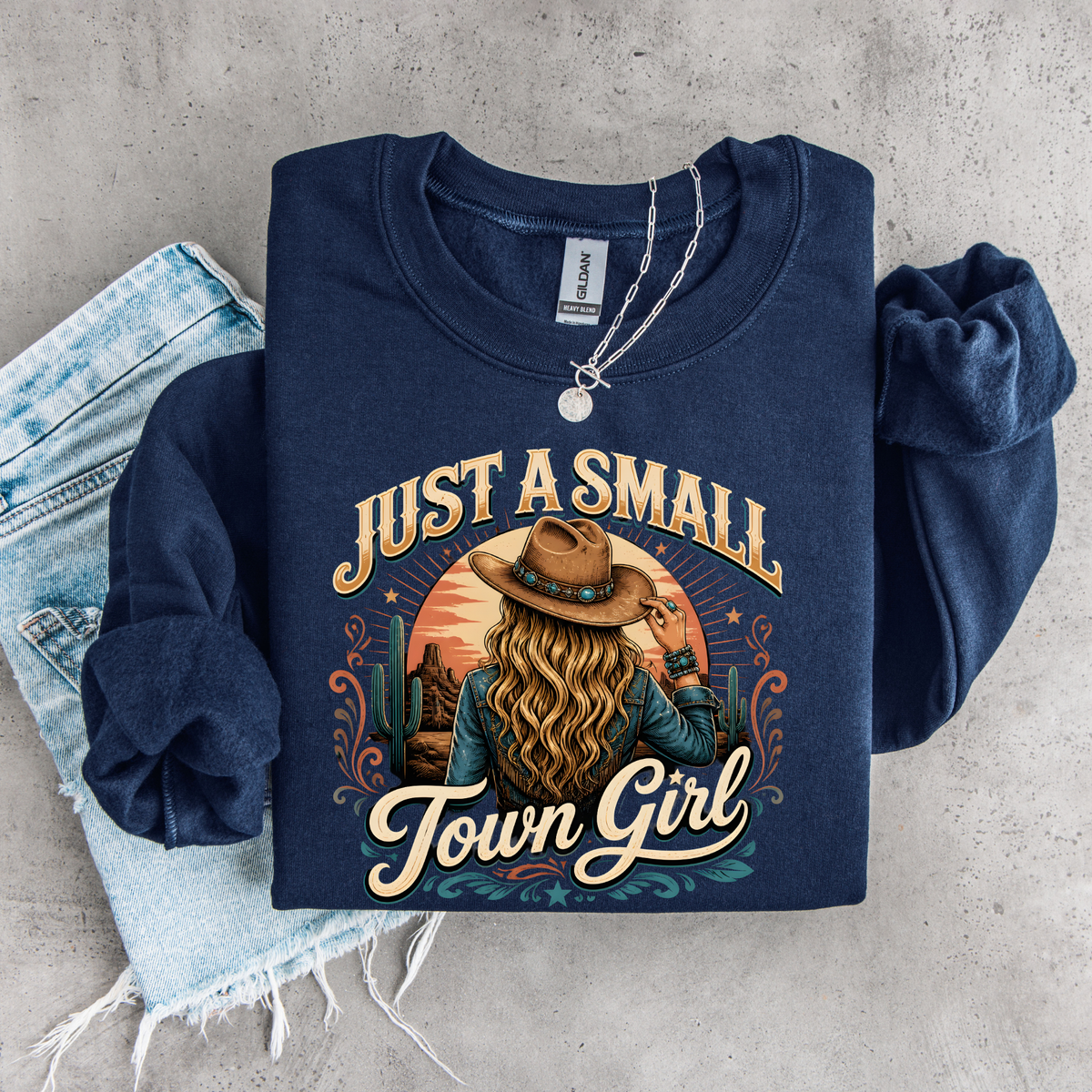 A navy blue long-sleeve crewneck sweatshirt with a graphic print of a woman in a cowboy hat and the text 'JUST A SMALL Town Girl', styled flat-lay on a gray concrete background with light-wash denim jeans and a silver necklace.
