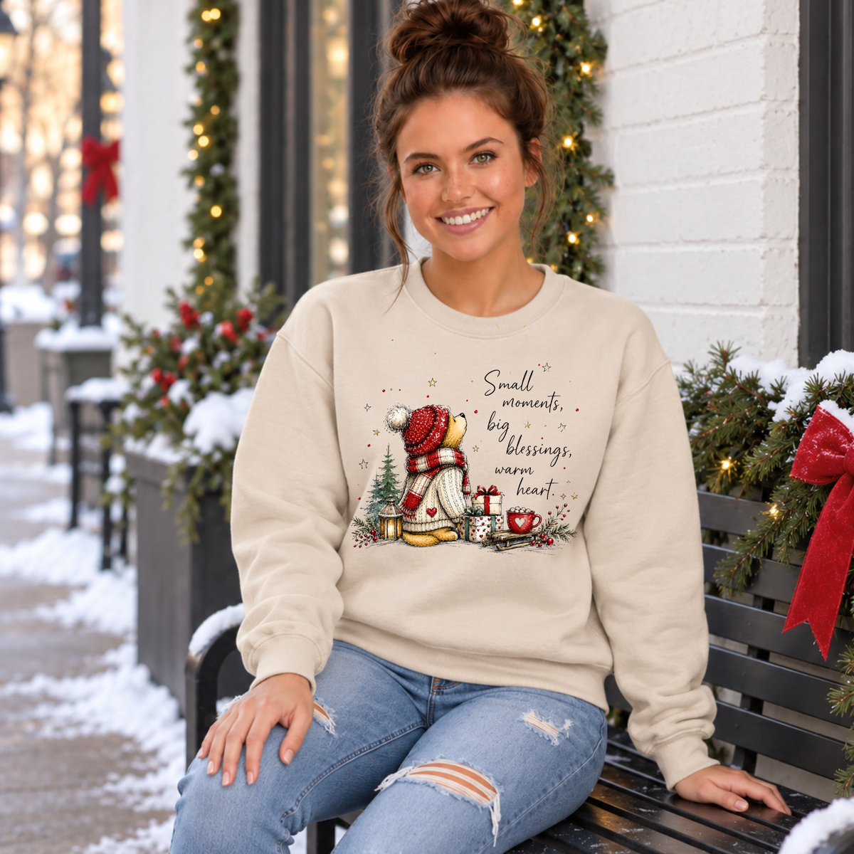 Woman seated on a snowy outdoor bench in a sand sweatshirt with winter bear and heartfelt holiday quote.