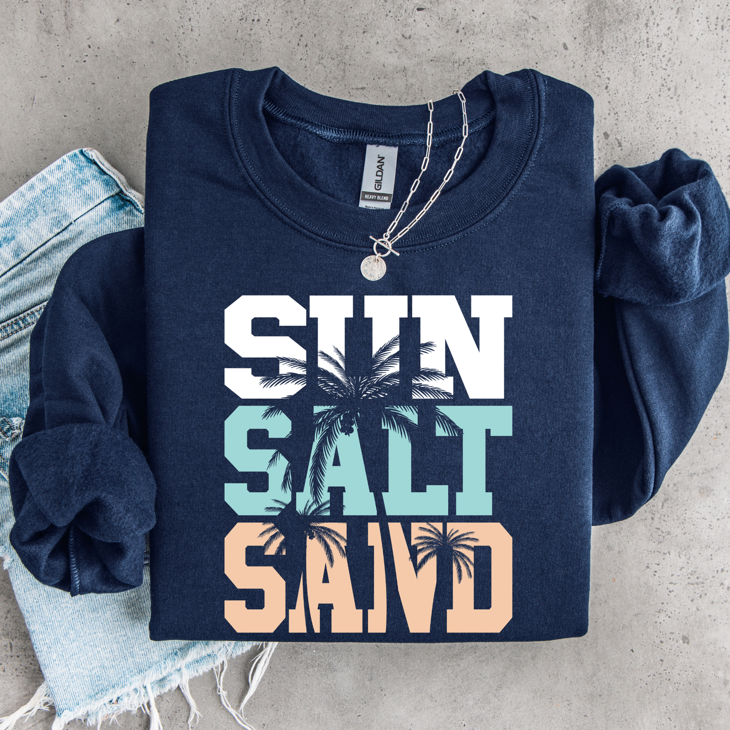 Folded navy blue sweatshirt with 'Sun Salt Sand' text and palm tree graphics on a concrete surface.
