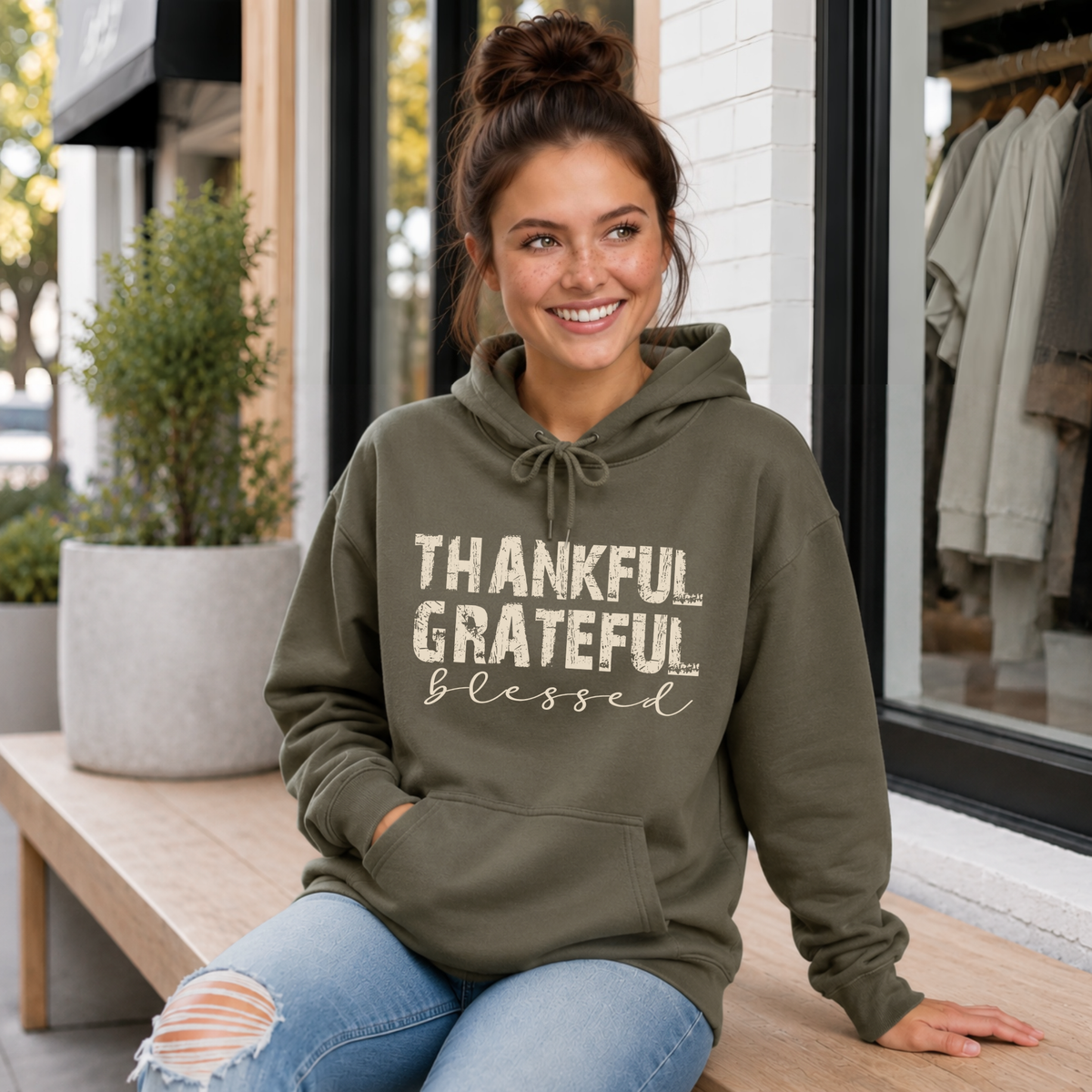 Woman seated in a military green hoodie with distressed Thankful Grateful Blessed text outside a boutique.