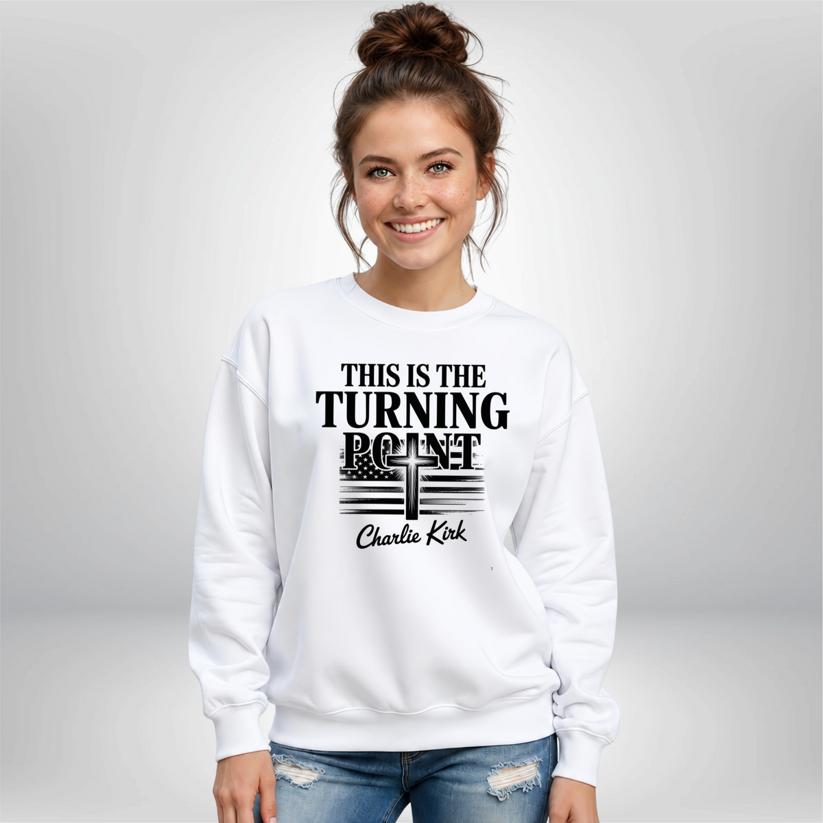 Woman against a gray background wearing a white crewneck sweatshirt with black This Is the Turning Point cross graphic.