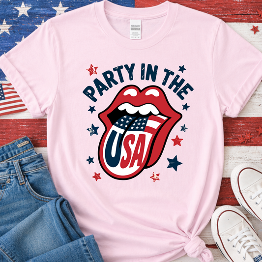 Pink t-shirt with 'Party in the USA' graphic on a wooden surface with American flags and jeans.