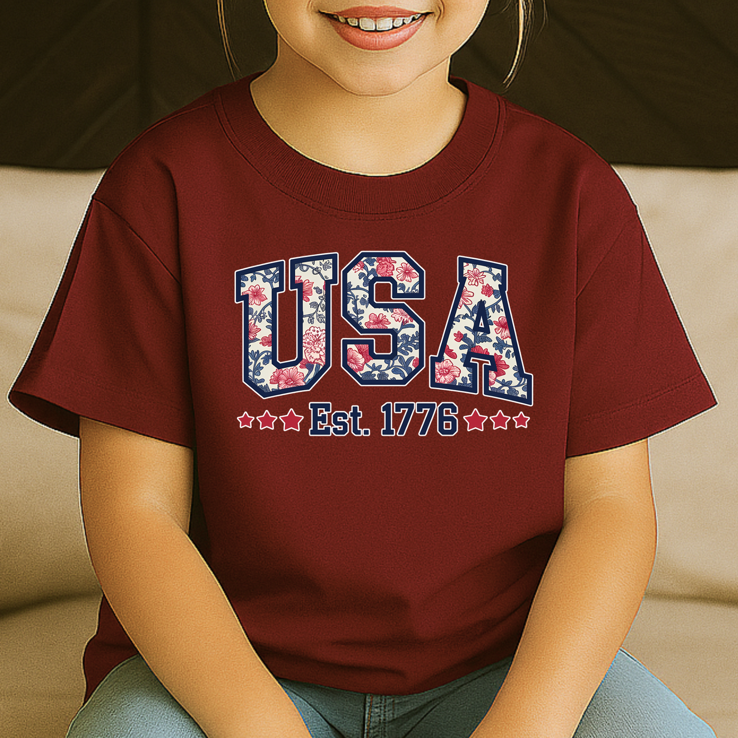 Youth girl in maroon USA Est. 1776 t-shirt, smiling while seated.