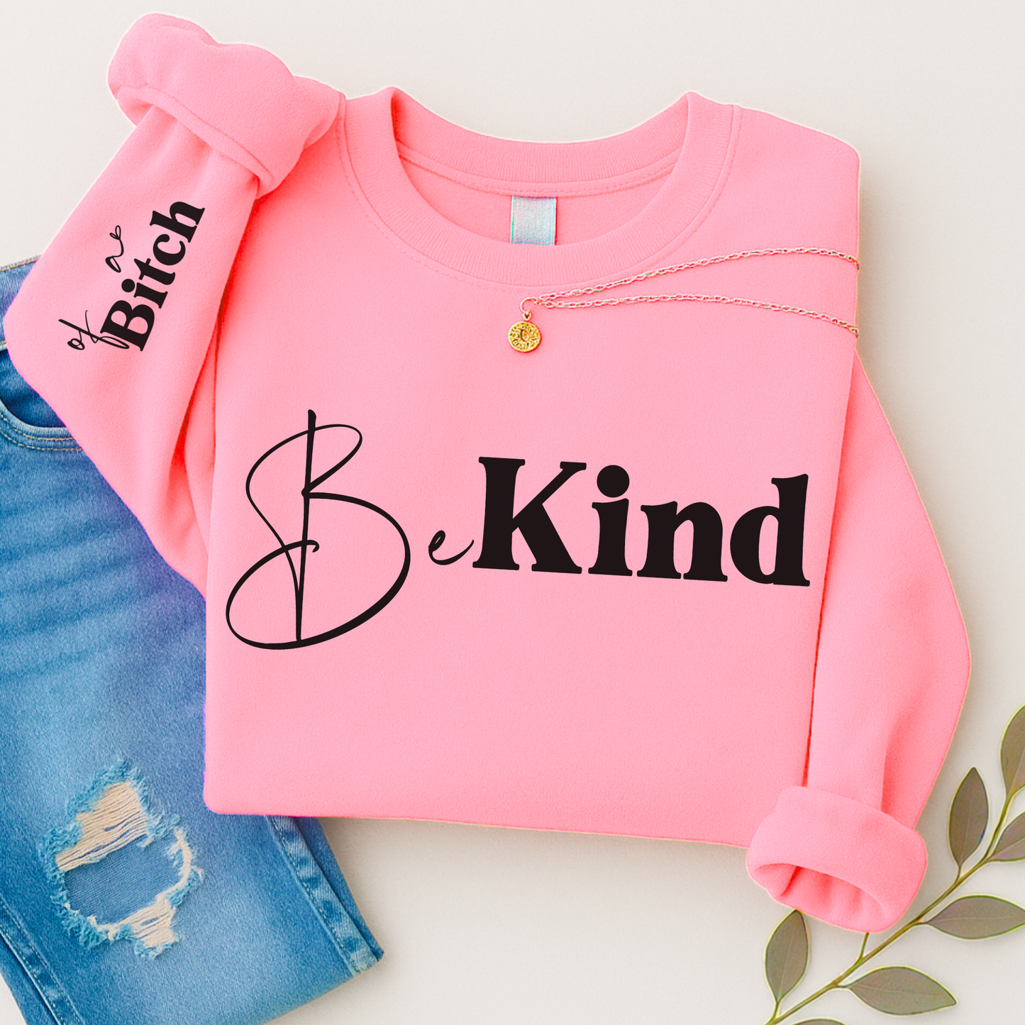 Pink sweatshirt with 'Be Kind' text, paired with denim jeans on a light background