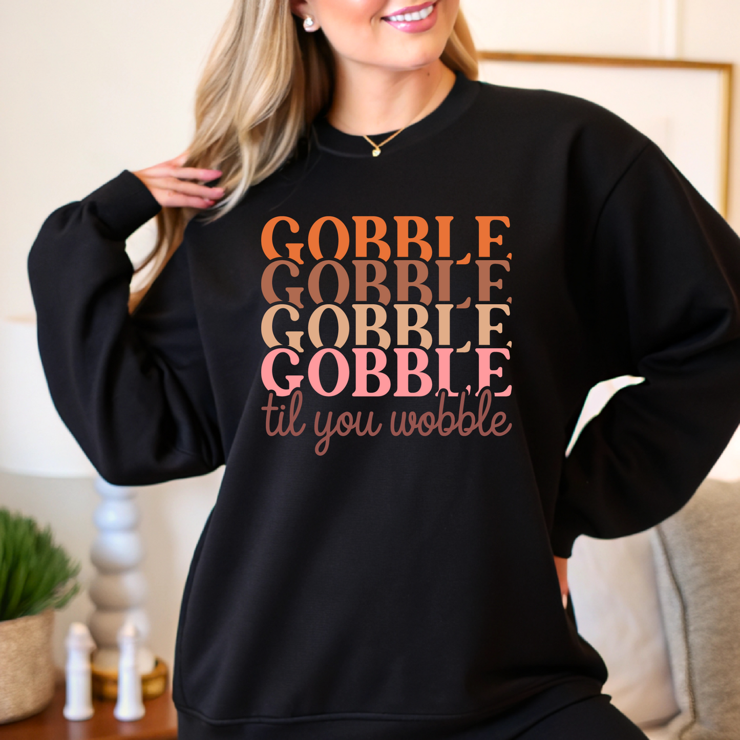 Black cozy fall sweatshirt with “Gobble Gobble” text in orange and pink tones – woman modeling in home setting.