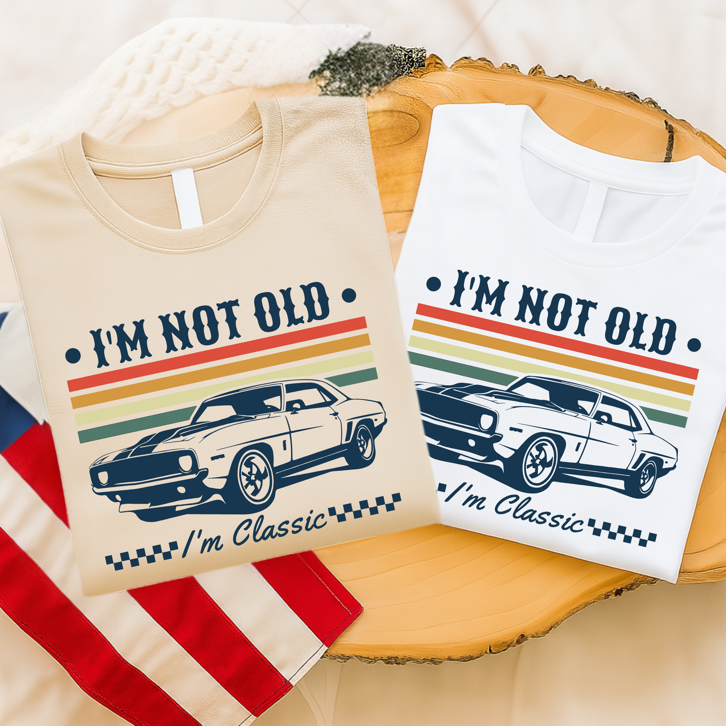 Flat lay of two folded T-shirts – one sand-colored and one white – both featuring the phrase “I’m Not Old – I’m Classic” with a retro car graphic and colorful stripes. Displayed on a wood table with patriotic American flag cloth. Ideal for Father's Day, birthdays, or car lovers.