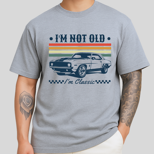 A man wearing a sport gray "I'm Not Old – I'm Classic" T-shirt featuring a retro muscle car and vintage stripe design. The shirt has a comfortable unisex fit and is styled for casual wear. Perfect for classic car lovers and guys with a timeless sense of humor.