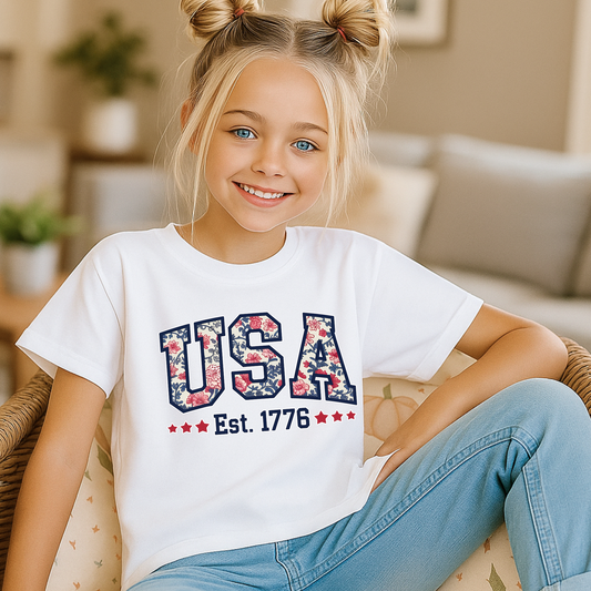 Girl relaxing in a white USA Est. 1776 tee, showing off red, white, and blue floral design.
