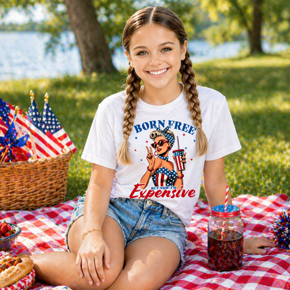 Girl sitting on a picnic blanket with a 'Born Free but Now Im Expensive' t-shirt, surrounded by American flags and a lake in the background.