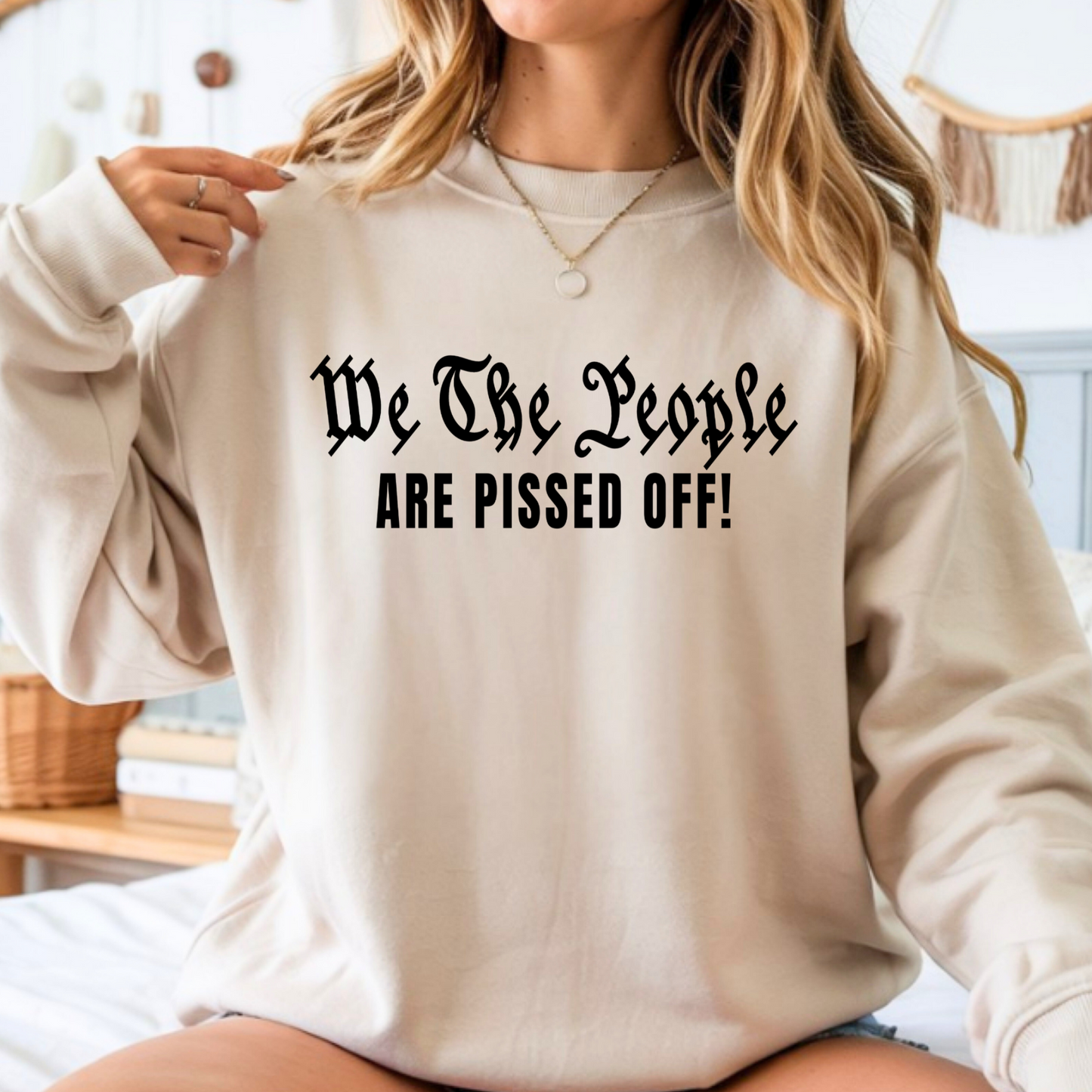 Person wearing a beige sweatshirt with text 'We The People ARE PISSED OFF!' in a casual setting.
