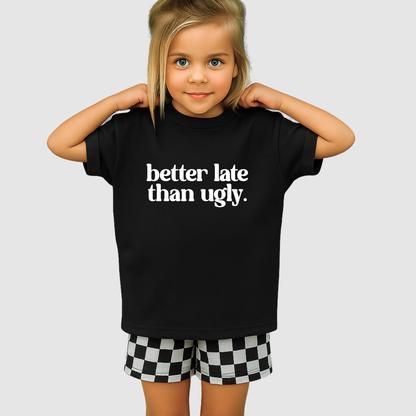 Child wearing a black t-shirt with 'better late than ugly' text and checkered shorts on a light gray background