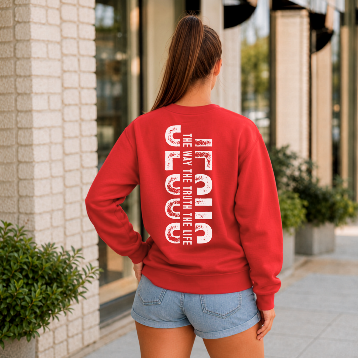 Woman shown from behind wearing a red crewneck sweatshirt with vertical white JESUS lettering and “The Way, The Truth, The Life” text.