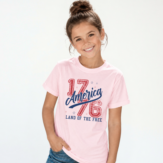 Smiling girl wearing a light pink youth T-shirt with a distressed red and blue America 1776 Land of the Free graphic