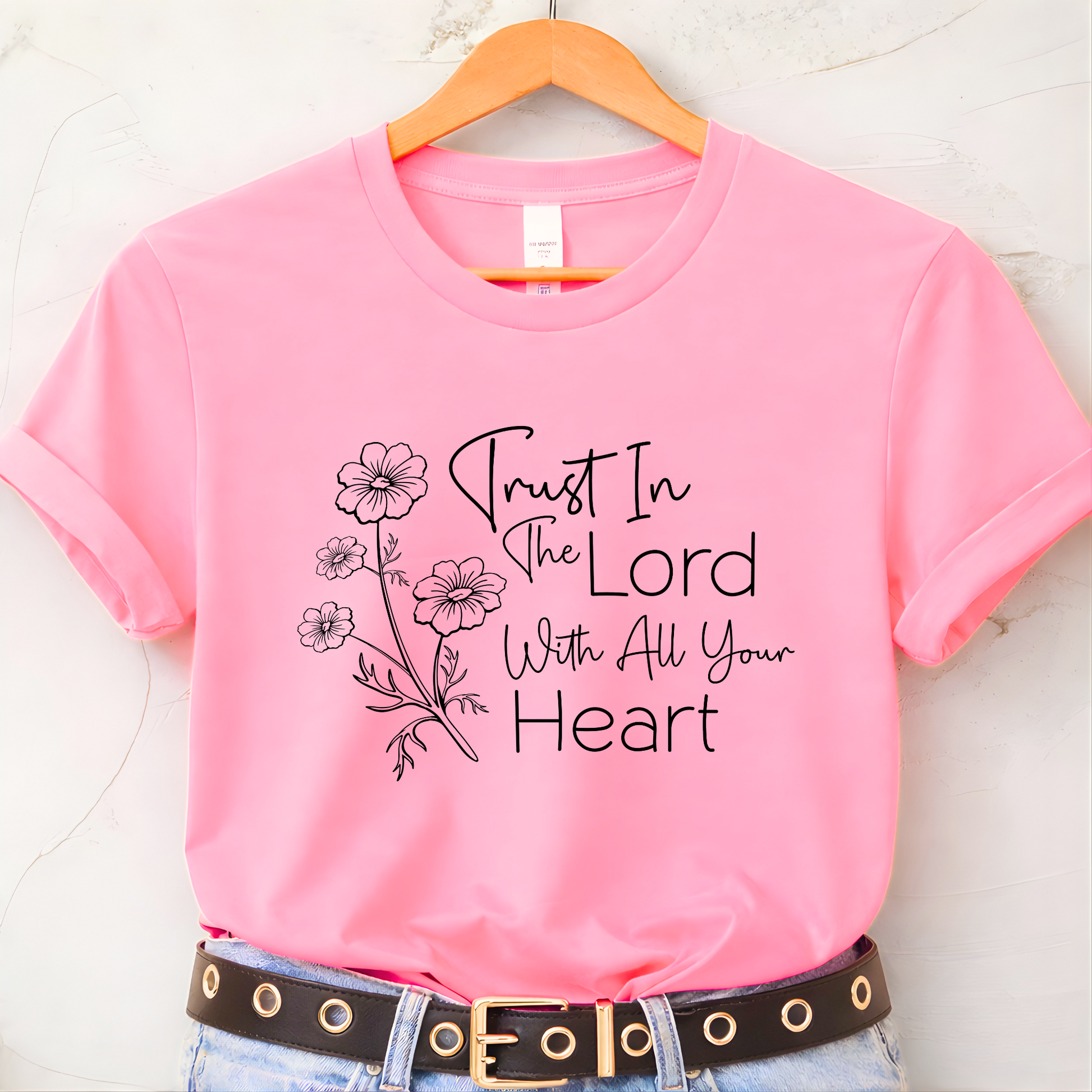 Pink t-shirt with 'Trust In The Lord With All Your Heart' text and floral design on a hanger.