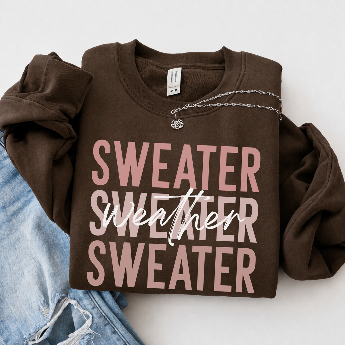 Folded brown crewneck sweatshirt with pink “Sweater Weather Sweater” graphic beside light-wash jeans on a neutral background.