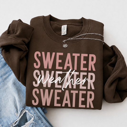 Folded brown crewneck sweatshirt with pink “Sweater Weather Sweater” graphic beside light-wash jeans on a neutral background.