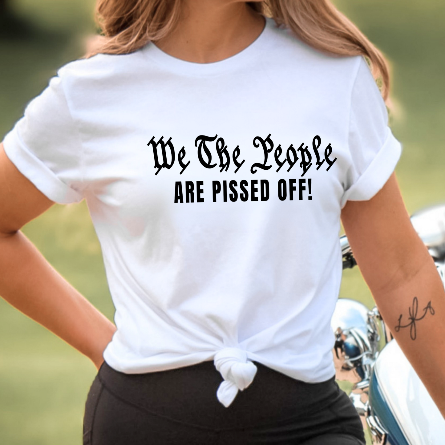 Woman wearing a white t-shirt with text 'We The People ARE PISSED OFF!' outdoors.