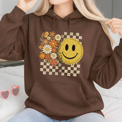 Woman wearing a brown hoodie with a smiley face and floral design, sitting on a bed.