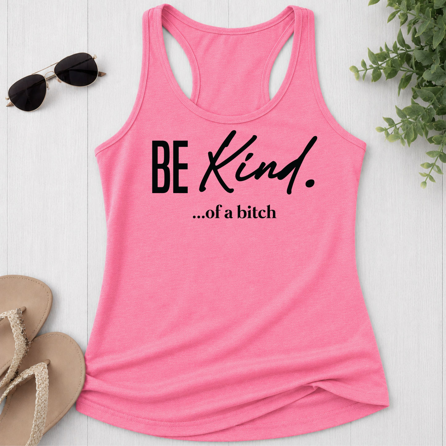 Pink tank top with 'BE Kind...of a bitch' text on a white surface with sunglasses and sandals.
