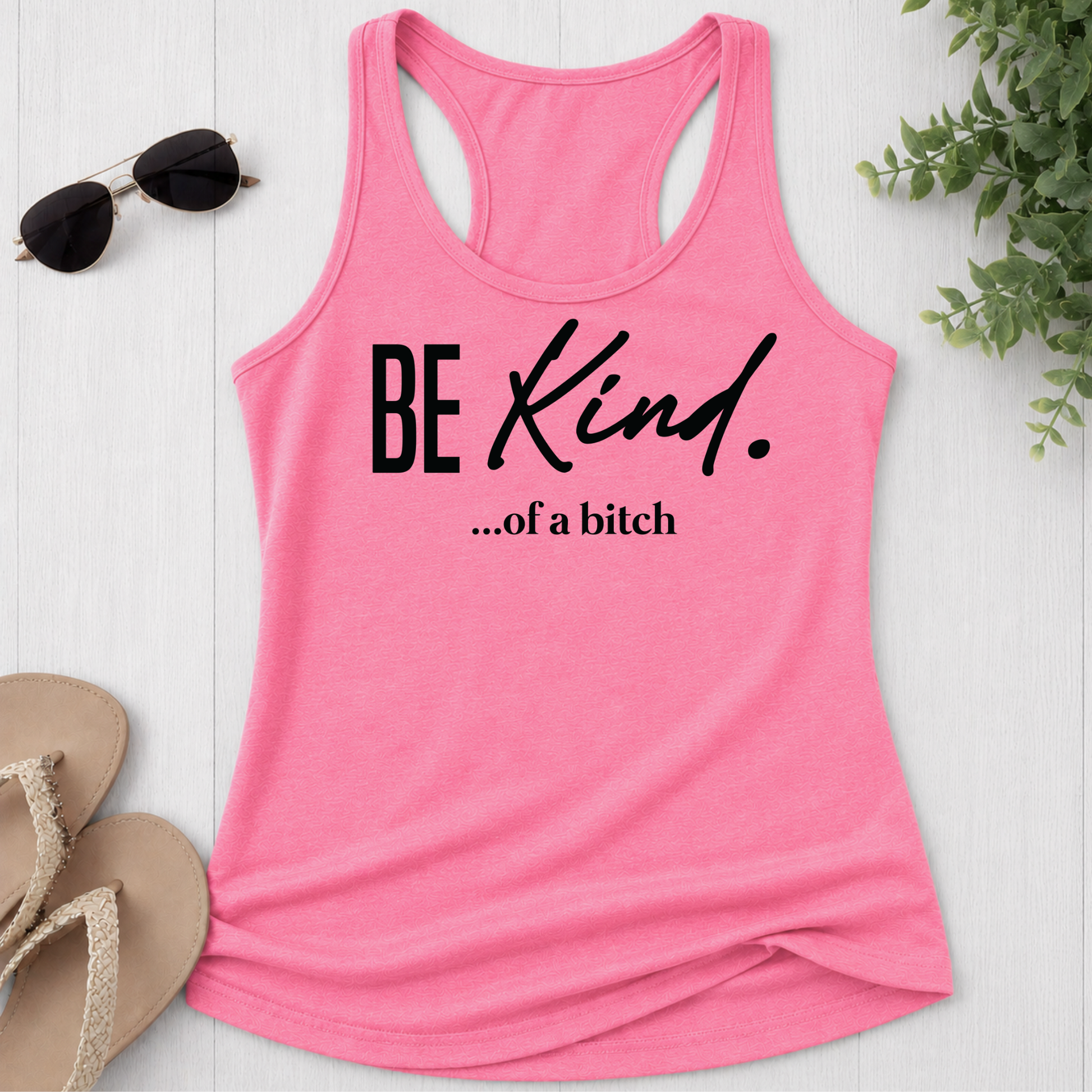 Pink tank top with 'BE Kind...of a bitch' text on a white surface with sunglasses and sandals.