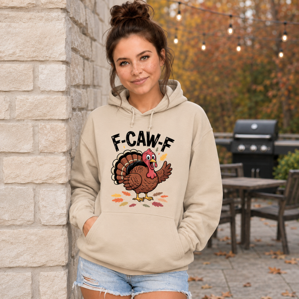 Woman wearing a sand hoodie with a colorful F-CAW-F turkey graphic in a fall patio setting.
