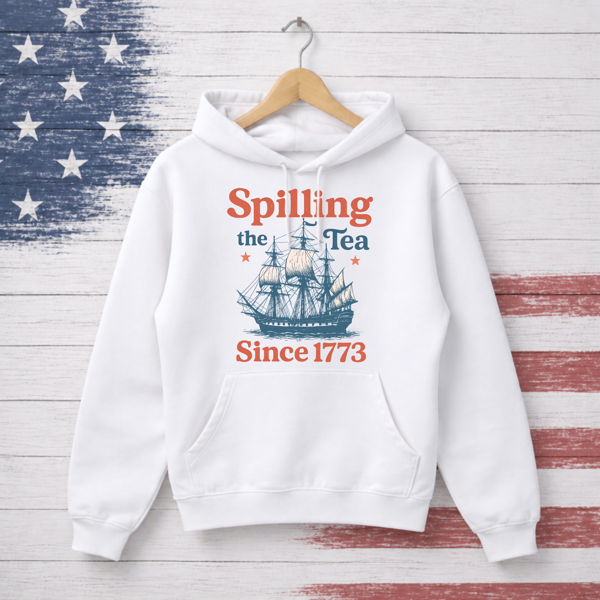 White hoodie with 'Spilling the Tea Since 1773' text and ship graphic on a wooden background.