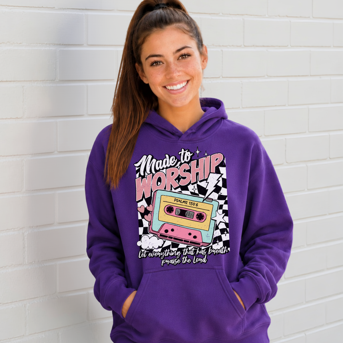 Woman wearing a purple Made to Worship hoodie with a retro cassette graphic against a white brick wall.