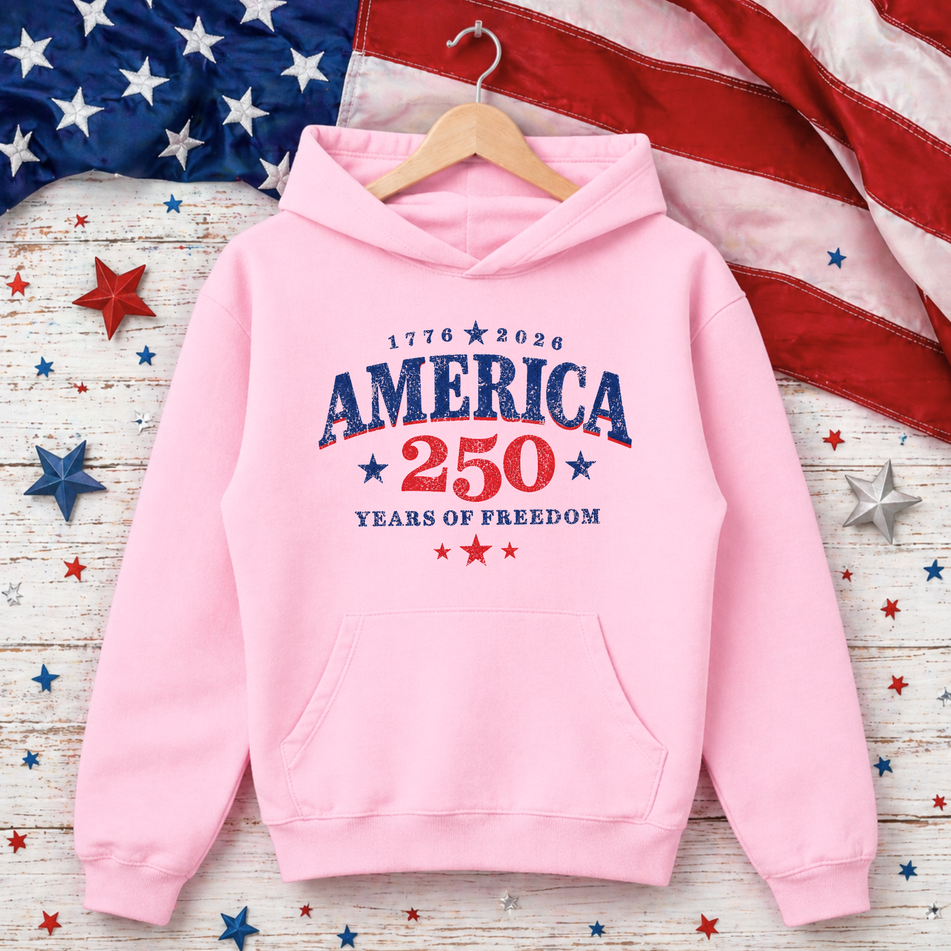 Pink hoodie with 'America 250 Years of Freedom' text on a wooden surface with American flag and stars.