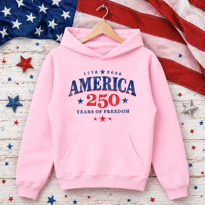 Pink hoodie with 'America 250 Years of Freedom' text on a wooden surface with American flag and stars.