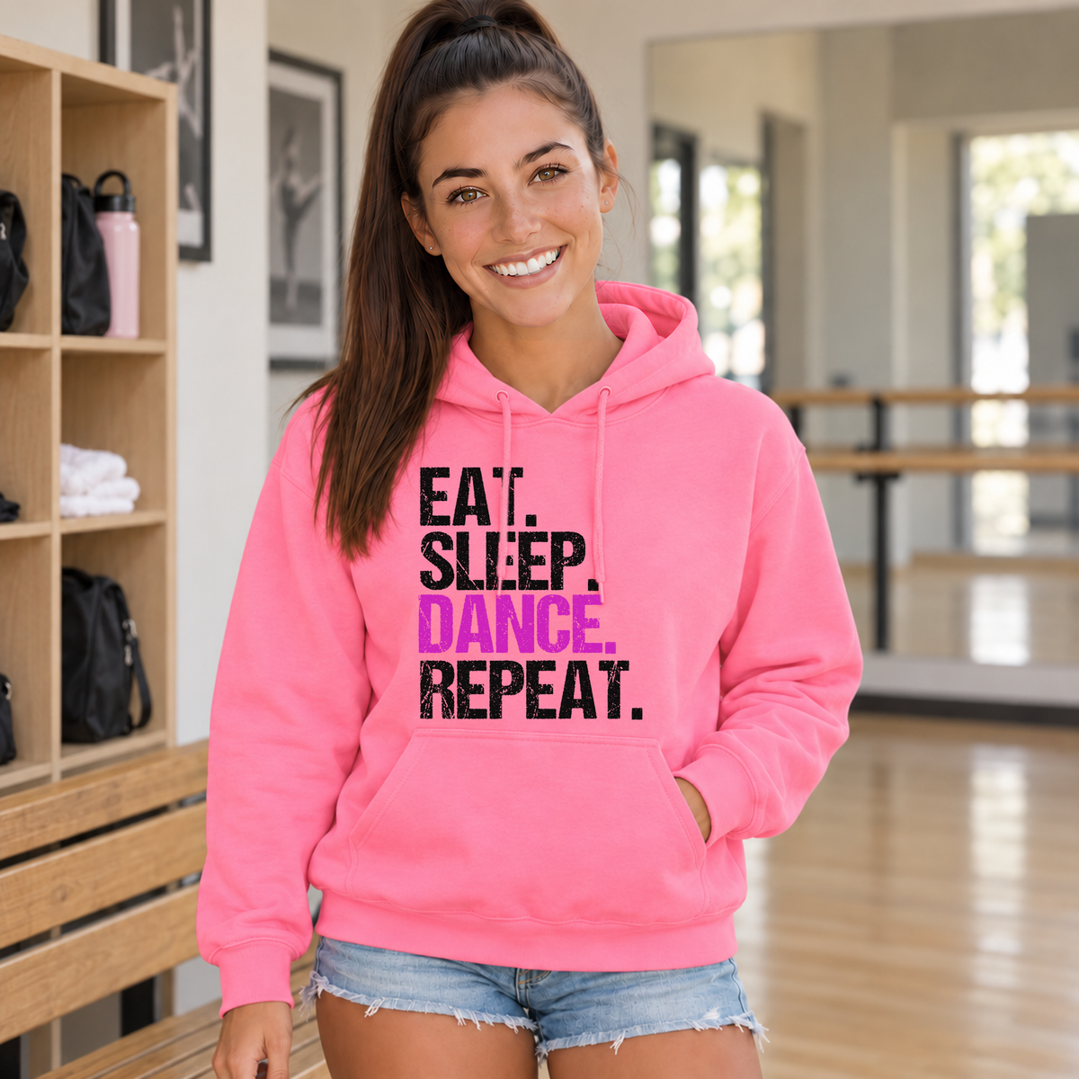 Woman wearing a Bright pink Eat Sleep Dance Repeat hoodie inside a dance studio with cubby shelves.