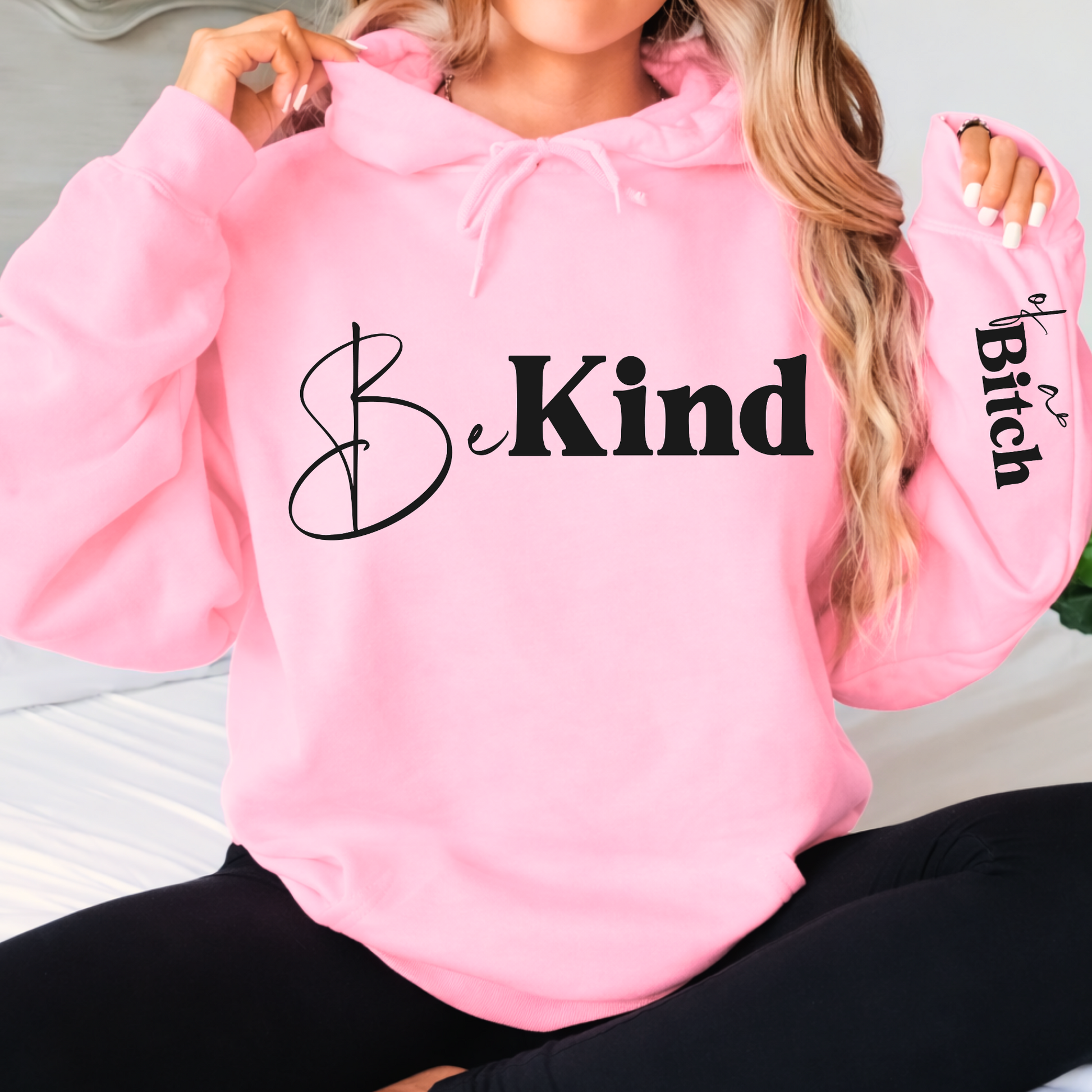 Pink hoodie with 'Be Kind' text on a person sitting on a white surface.