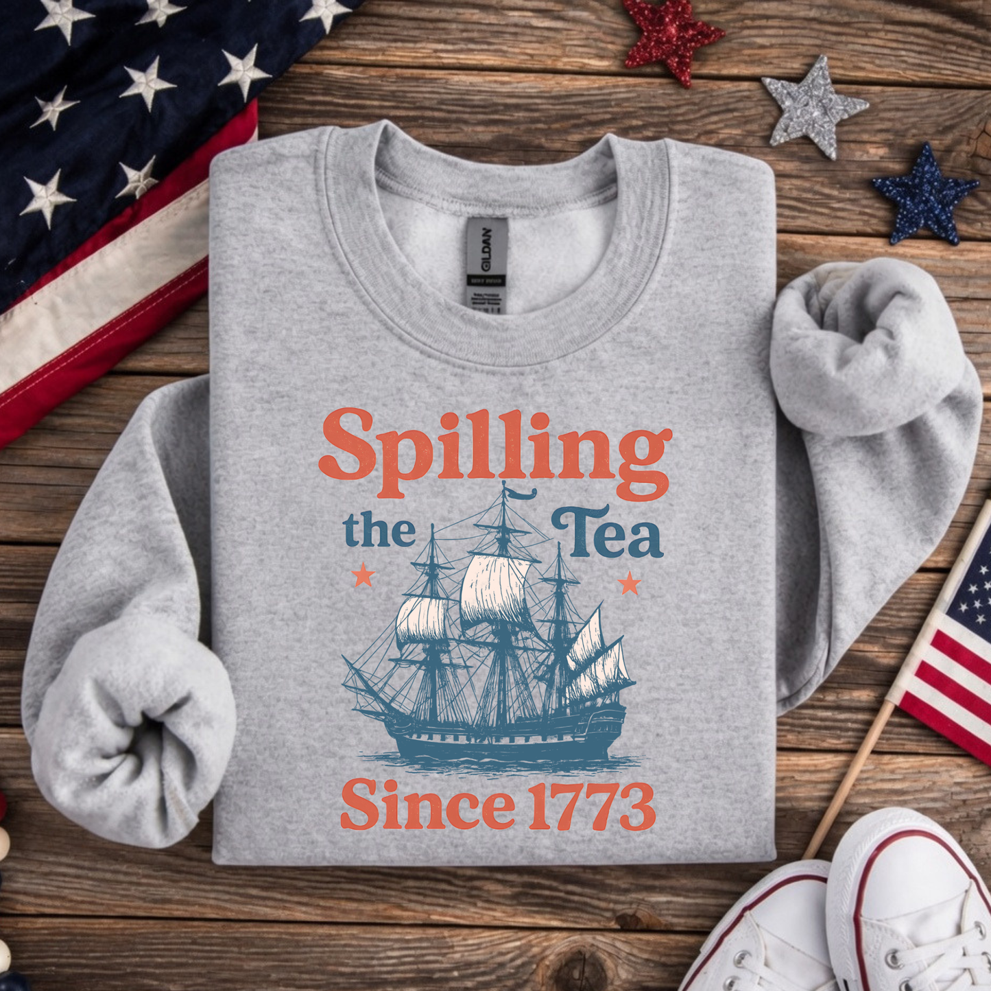 Gray sweatshirt with 'Spilling the Tea Since 1773' design on a wooden surface with American flags.