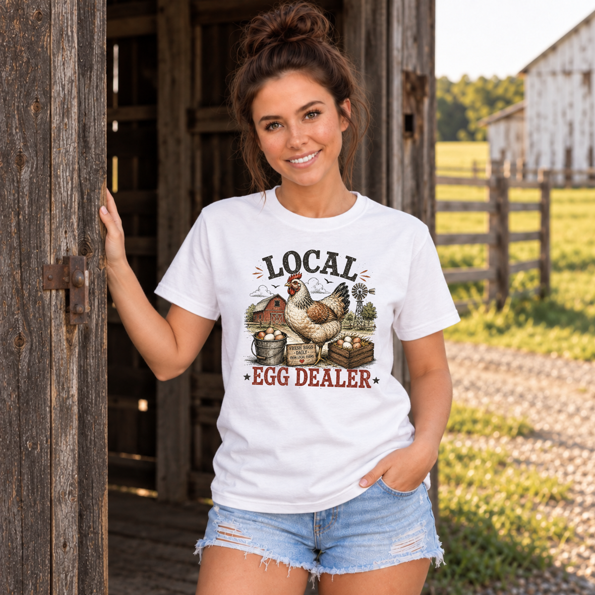 Woman in a white Local Egg Dealer T-shirt standing by a barn door with a fenced pasture behind her.